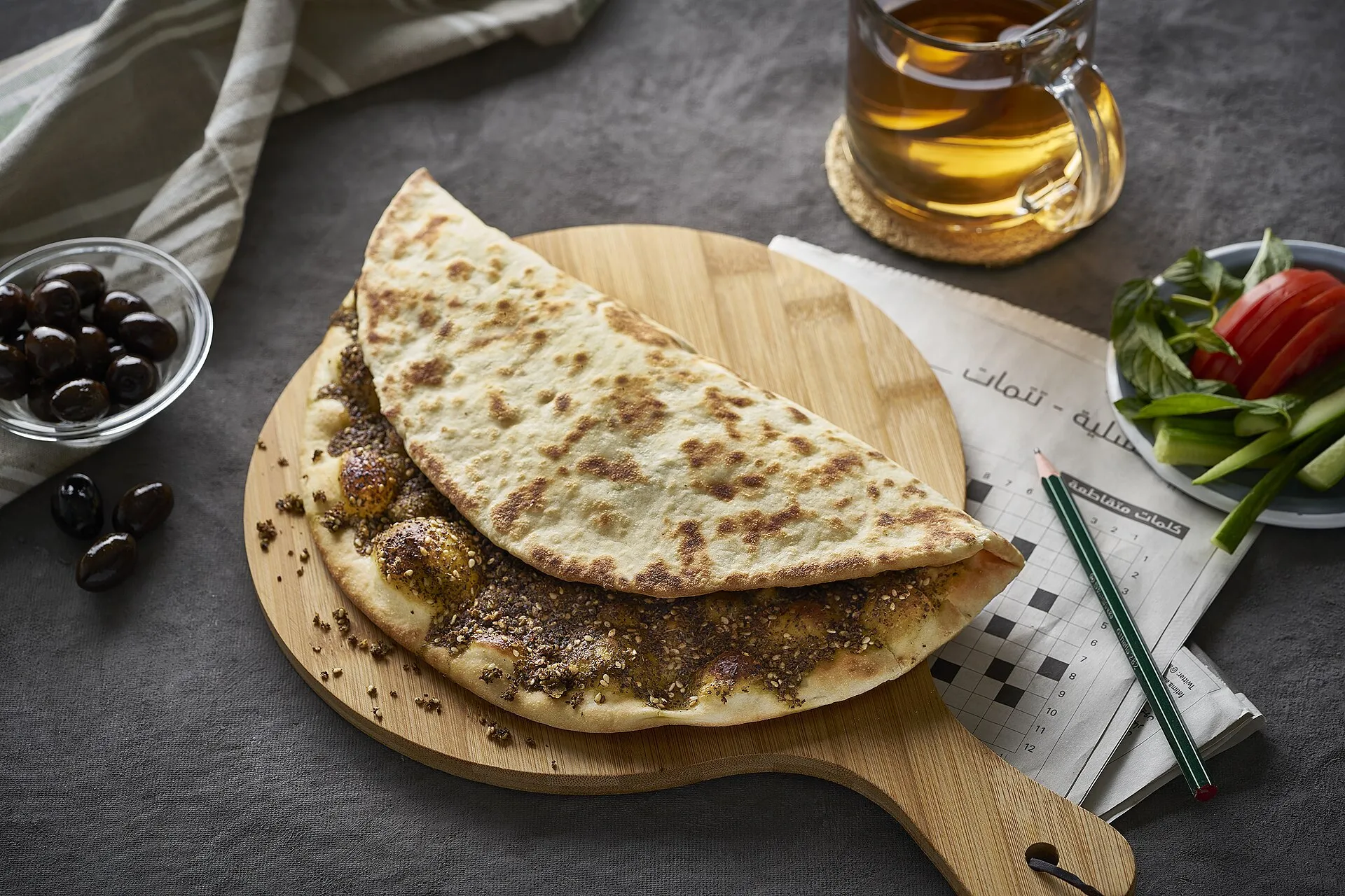 Manakish mit Za'atar auf Holzbrett, daneben Oliven und Tee